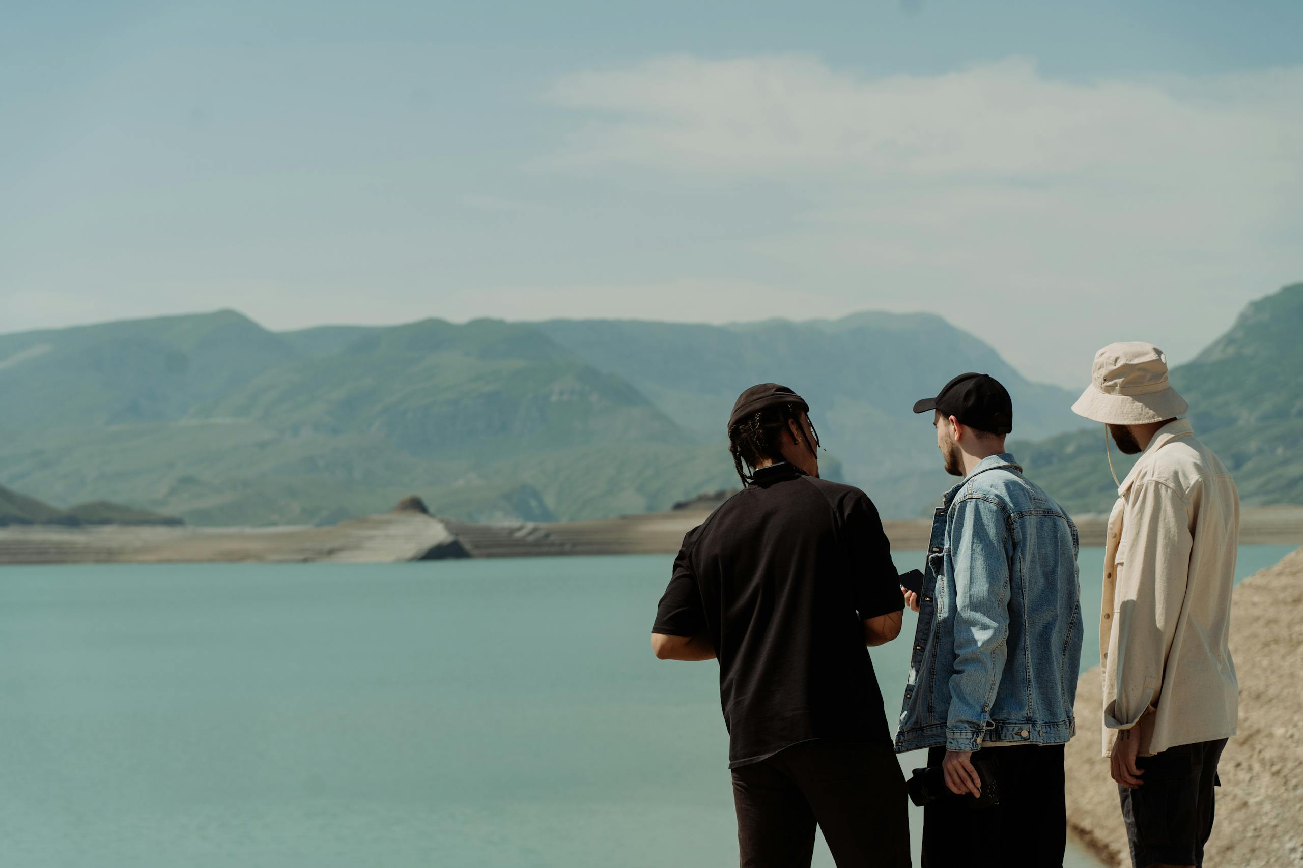 Three men in hats stand by a serene mountain lake, enjoying the scenic landscape under a clear sky.